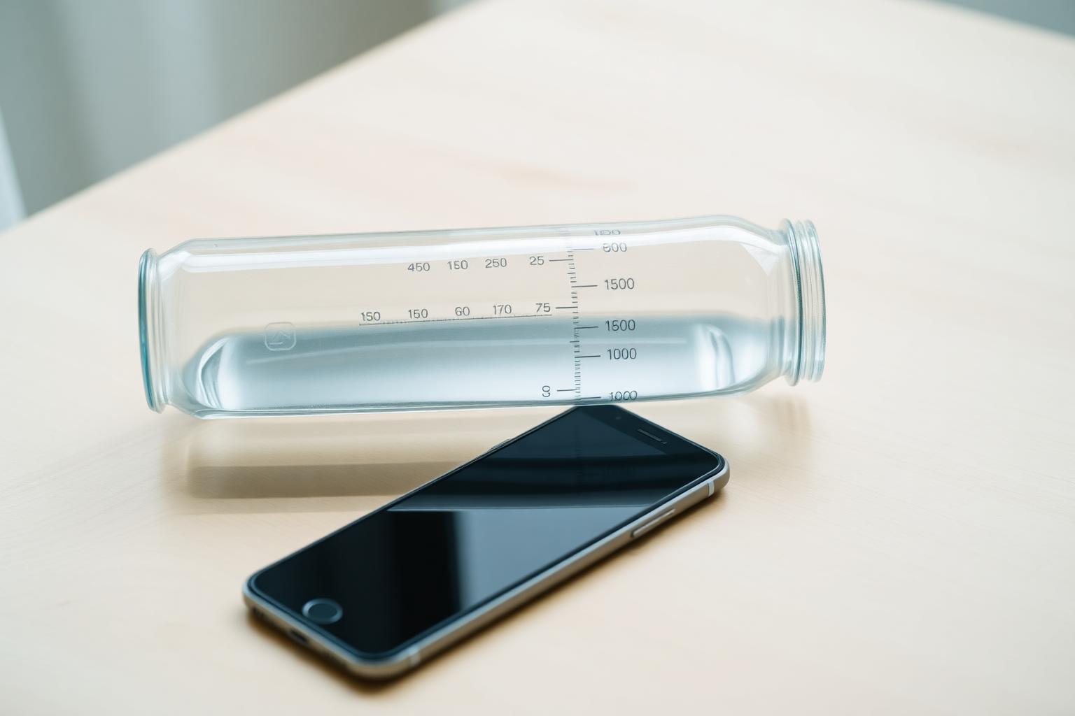 Reusable glass water bottle with measurement markings next to a smartphone showing a hydration tracking app on a light wooden desk — using a water intake calculator